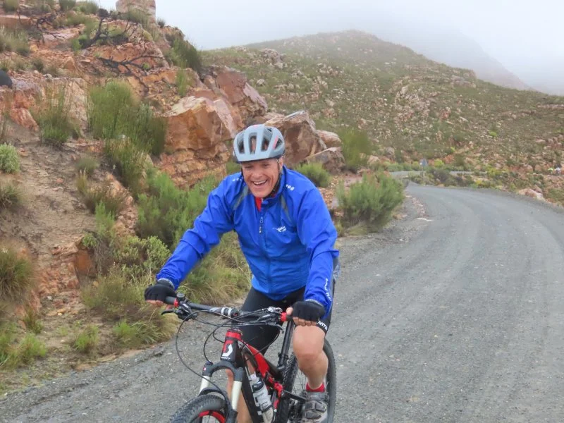 Person smiling and riding a mountain bike on a gravel trail in a mountainous area with shrubs and rocks.