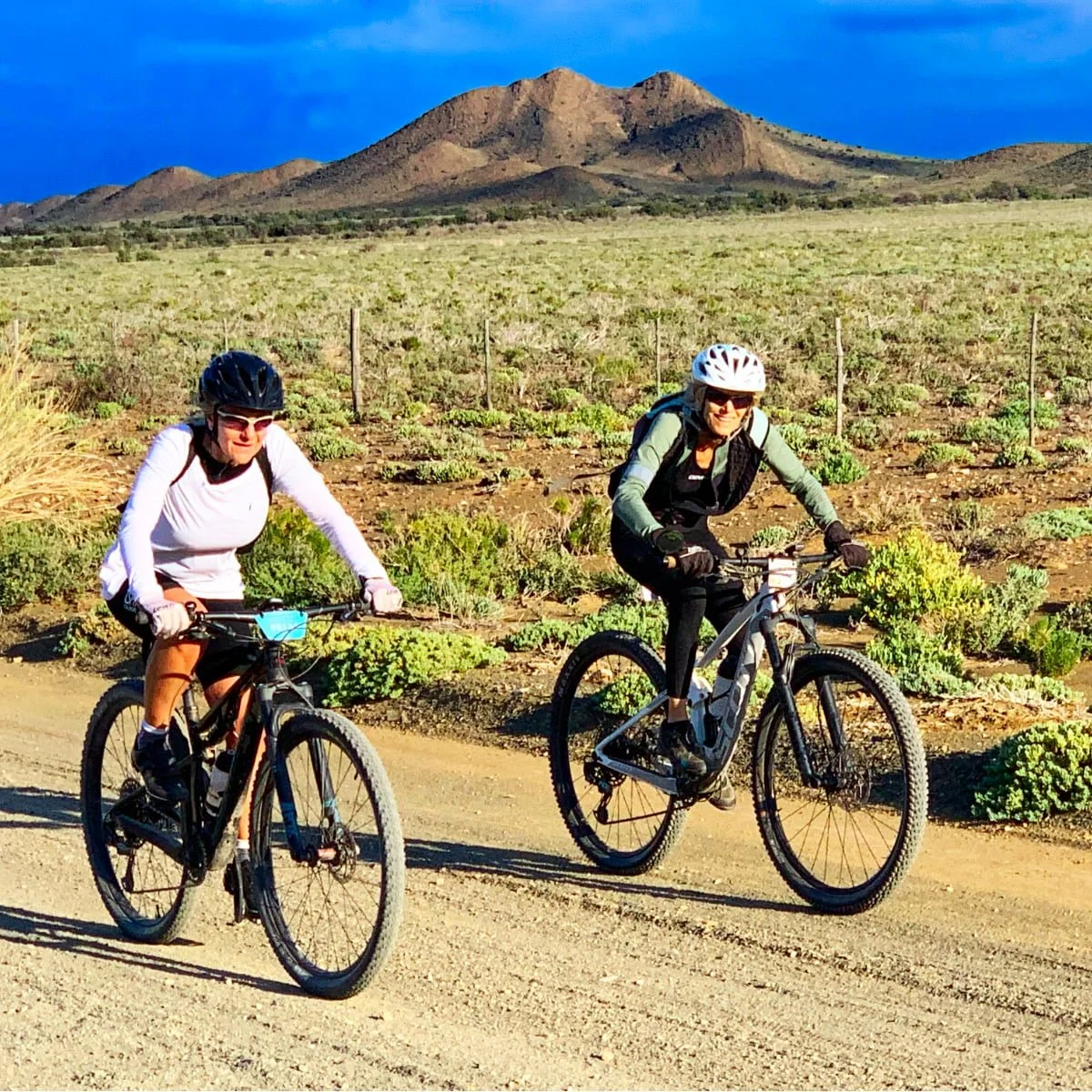 Two women riding mountain bikes on a dirt trail in a desert landscape with mountains in the background. They are wearing helmets and sunglasses, smiling as they enjoy a sunny day.