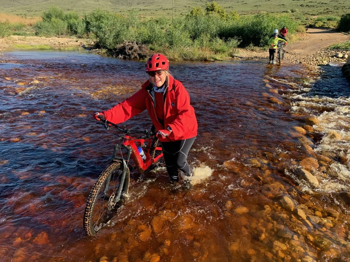 Woman in a red jacket and helmet standing with a mountain bike in a shallow creek, with other people riding bikes in the background on a trail.