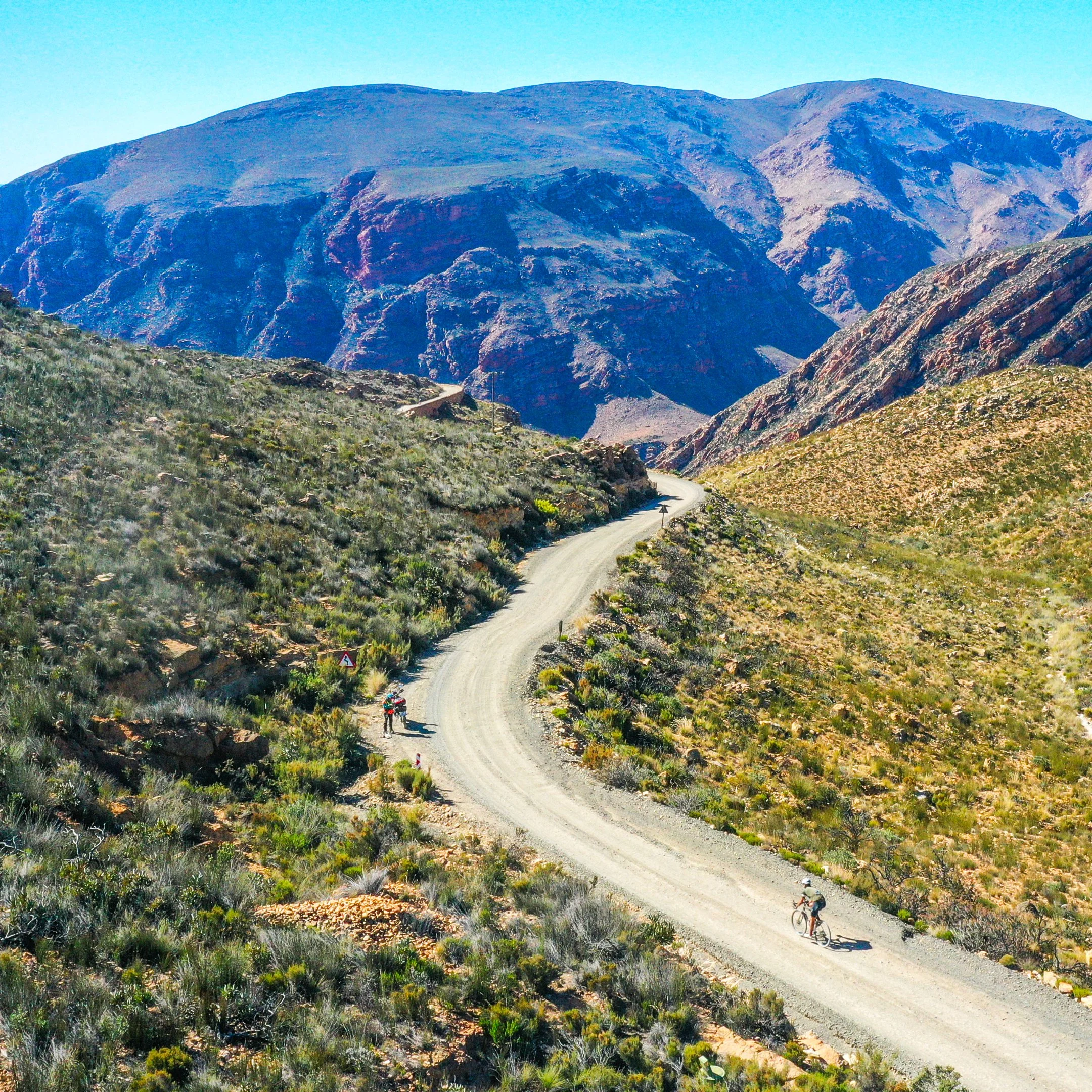 A winding dirt road through a mountainous desert landscape with two cyclists riding along it.