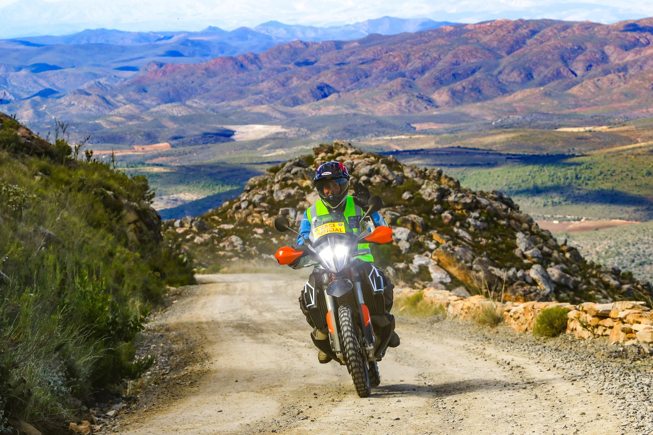 A motorcyclist in a high-visibility vest and helmet riding on a dirt road through a mountainous landscape, with colorful hills and valleys in the background.