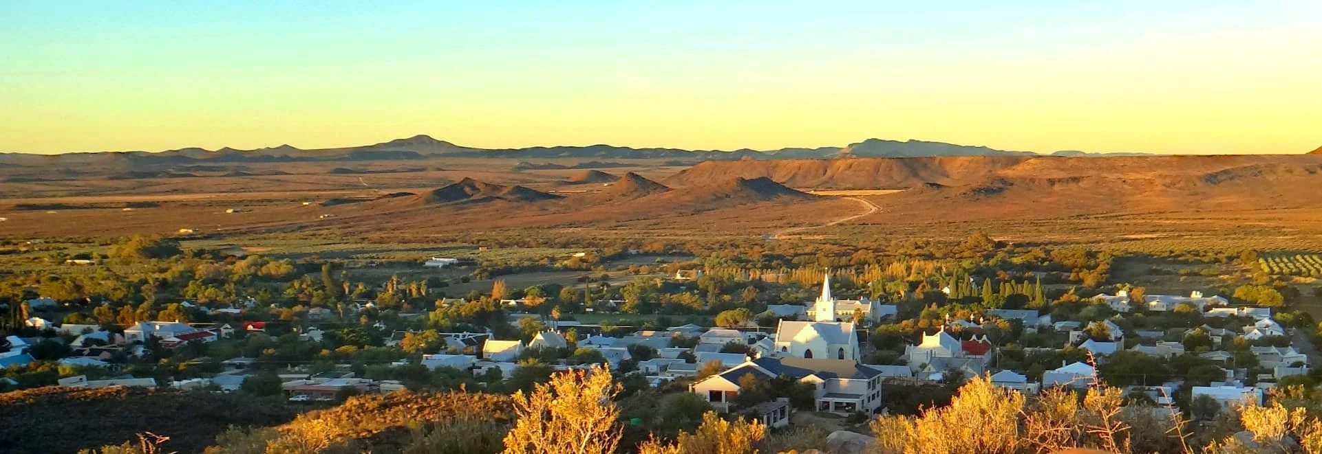 A small town with white buildings, including a church with a steeple, surrounded by trees and mountains in the background during sunset.