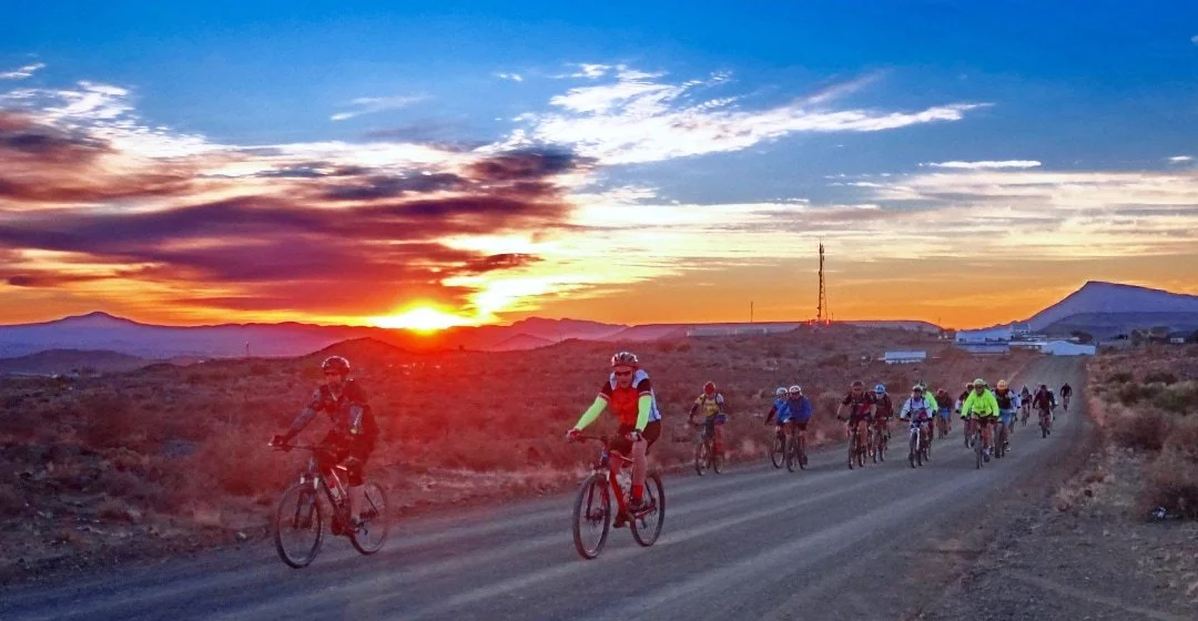 Group of cyclists riding on a dirt road during sunset in a desert landscape with mountains in the background.