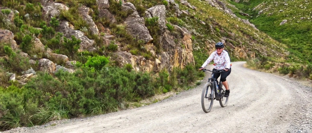 Woman riding a mountain bike on a gravel trail through a hilly, rocky landscape with green vegetation.