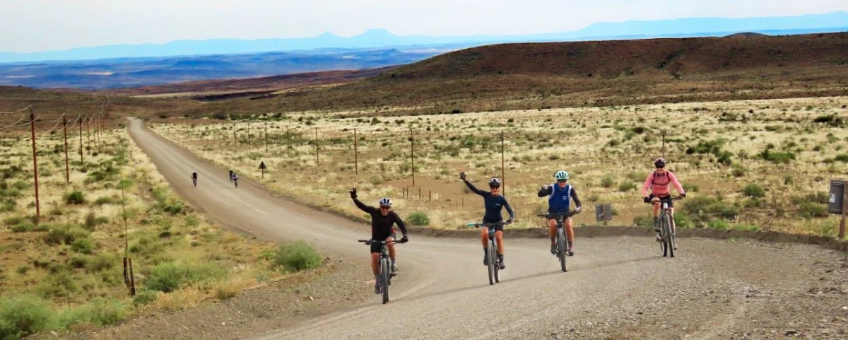 Group of cyclists riding on a dirt road through a desert landscape with sparse vegetation, utility poles, and mountains in the background.