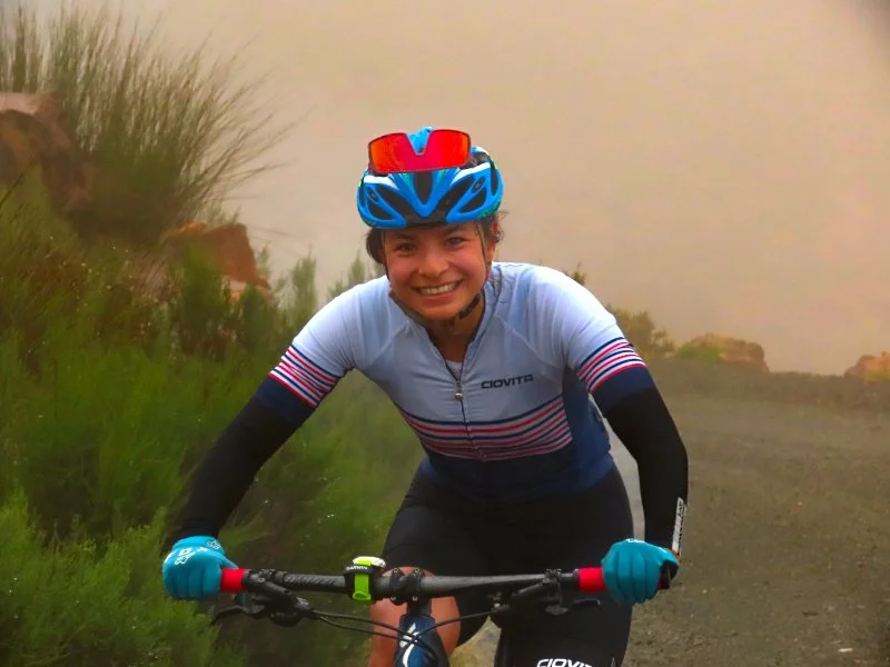 Smiling woman mountain biking in a foggy outdoor setting with green vegetation and a dirt path.