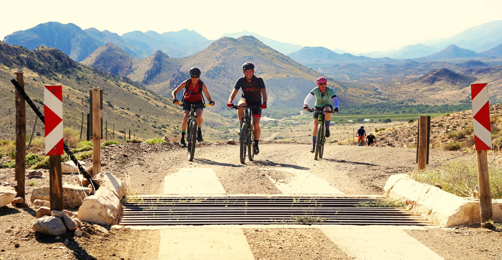 Four cyclists riding mountain bikes on a dirt trail in a mountainous landscape with distant peaks, some of the trail segments are blocked with a man-made barrier.