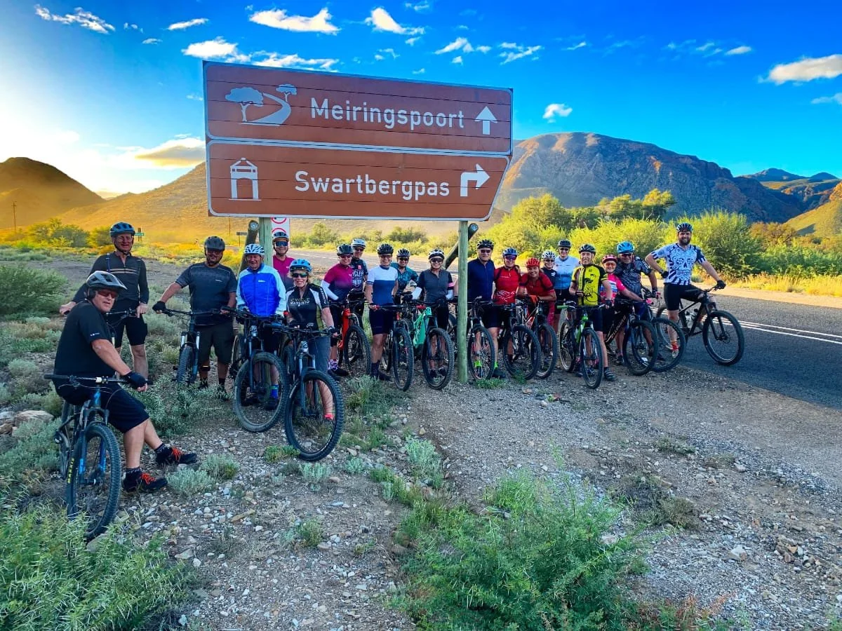 Group of cyclists standing next to a road sign in a mountainous area with clear blue sky, green bushes, and mountains in the background.