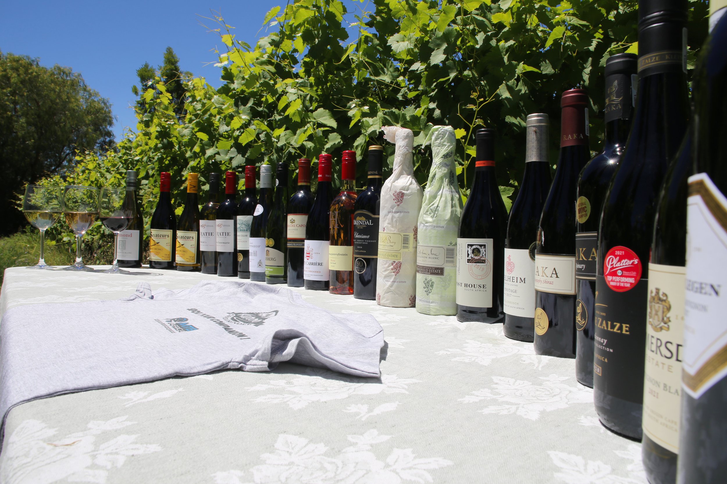 A table outdoors with various bottles of wine, some wrapped, and two glasses of white wine, set against a background of green grapevines and a bright blue sky.