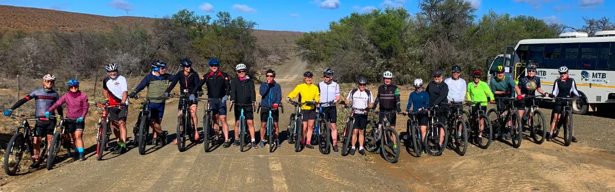 Group of people with mountain bikes standing on a dirt trail outdoors, with a bus in the background and a partly cloudy sky.