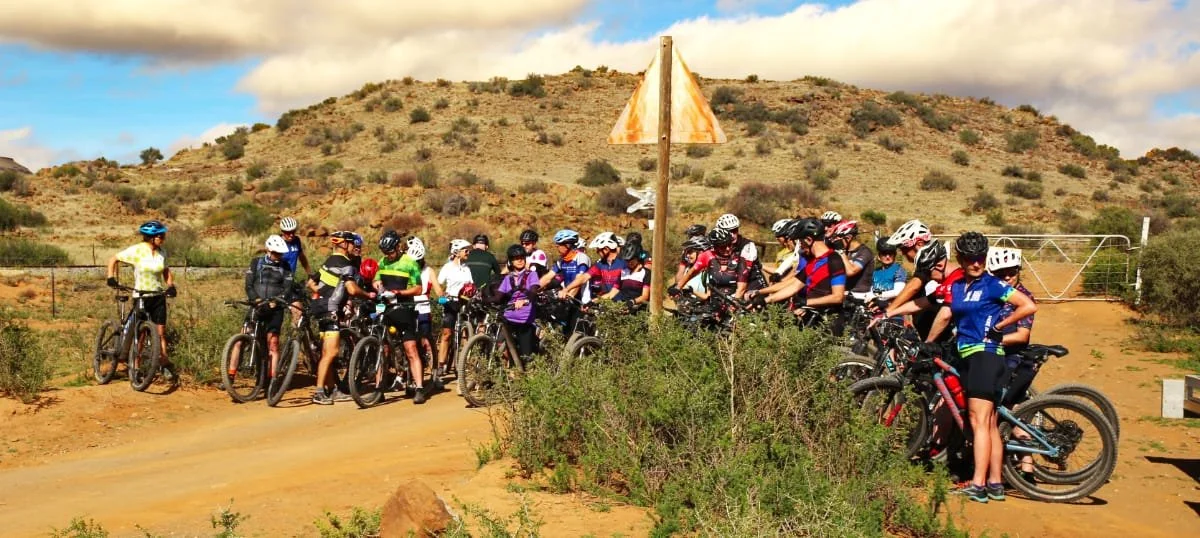 Group of children and teenagers with bikes, wearing helmets, gathered at a trailhead in a desert landscape, possibly preparing for a bike ride.