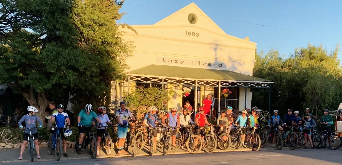 Group of people with bicycles standing in front of the Lazy Lizard building, which has the date 1903 on it, under a porch with hanging plants, during daytime.