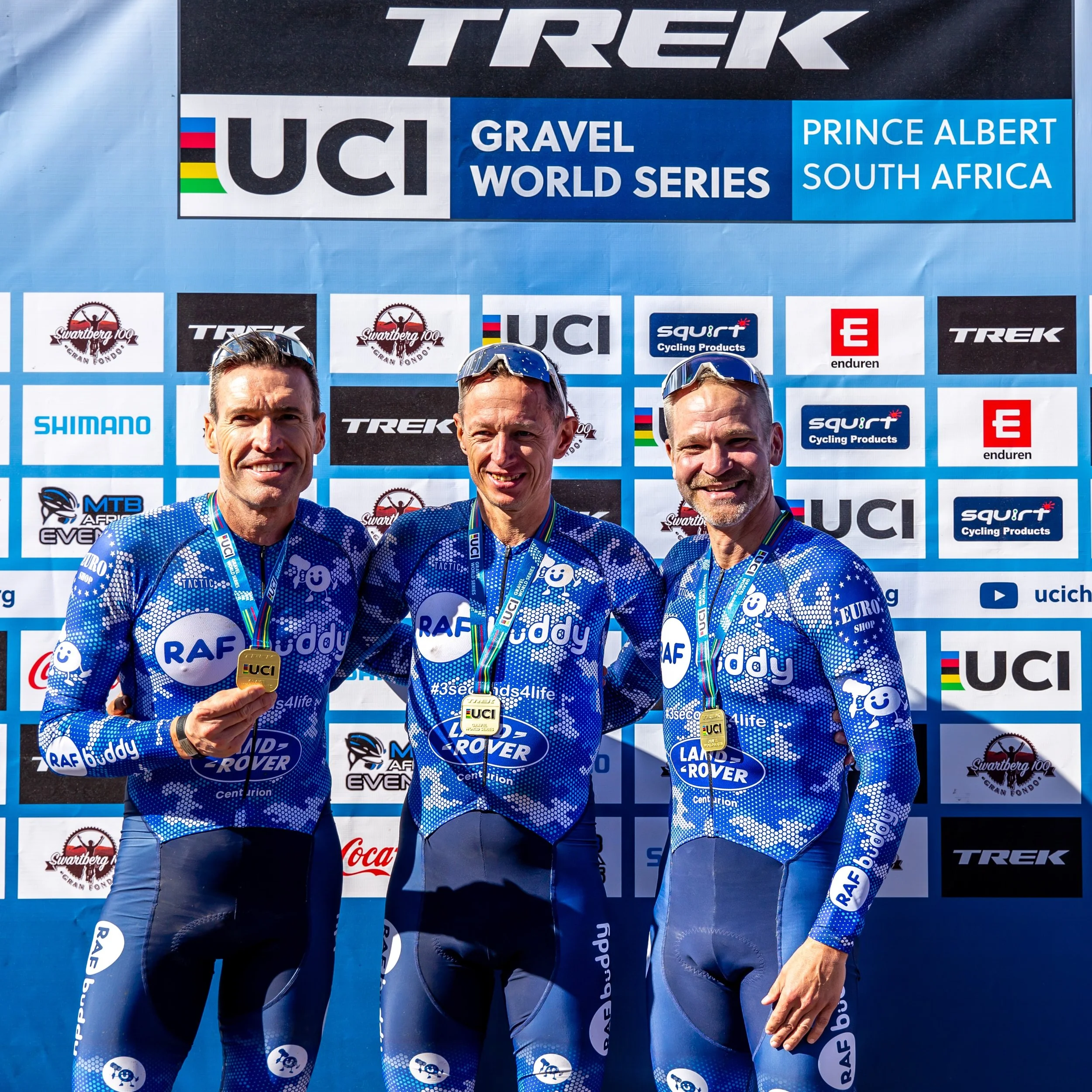 Three male cyclists in blue Team RAF jerseys standing on a podium, each wearing medals, celebrating their achievement at a UCI gravel cycling event in South Africa.