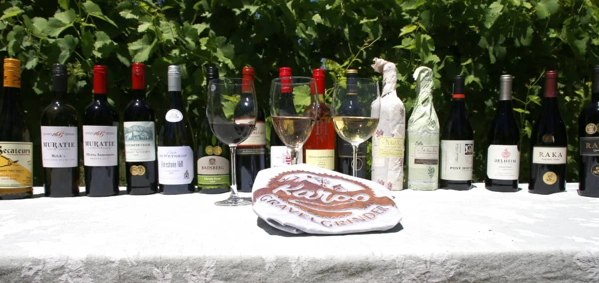 A row of various wine bottles and two glasses of wine on a white tablecloth outdoors, with green foliage in the background.