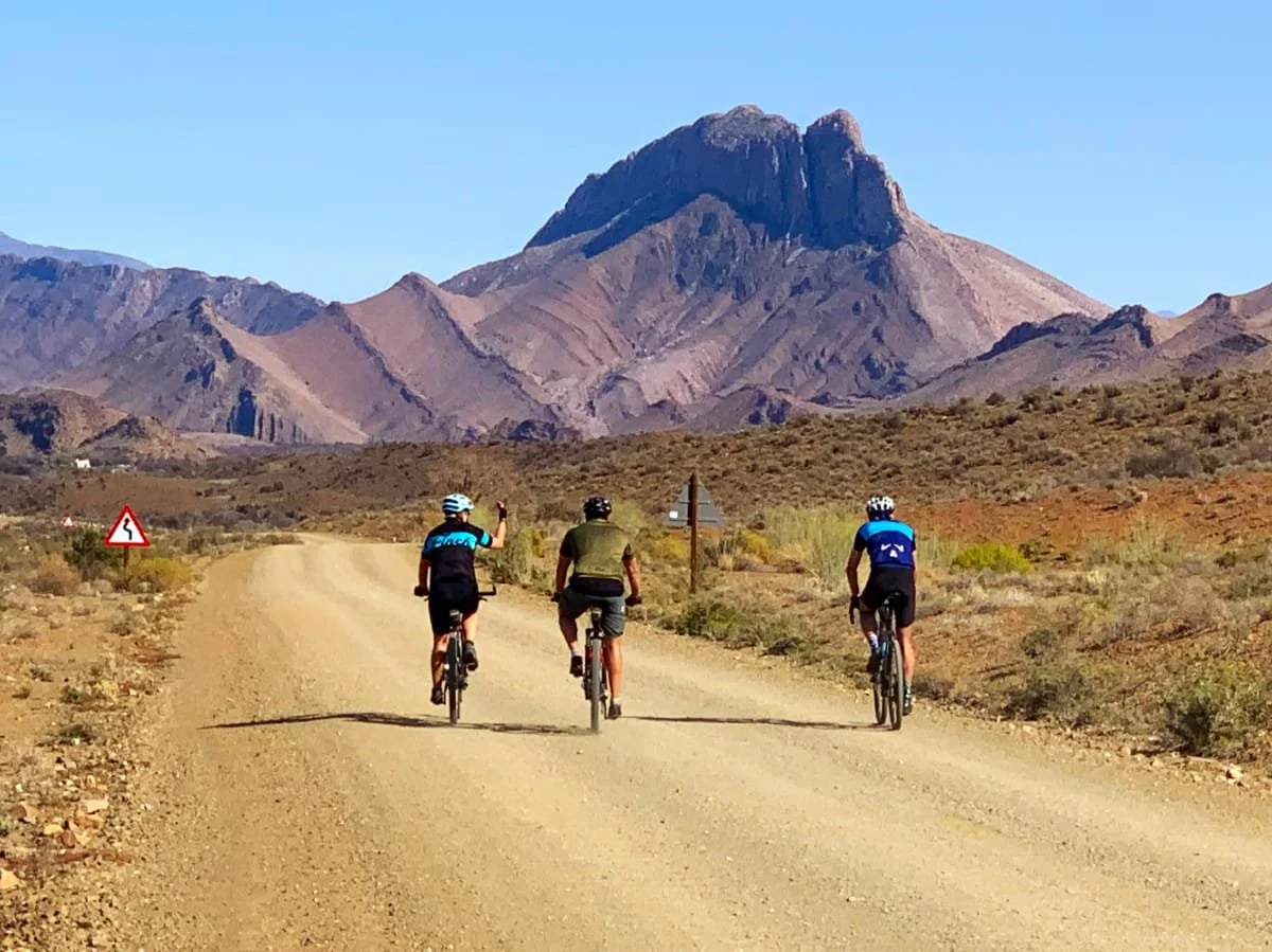 Three bikers riding on a dirt road through a desert landscape with mountains in the background.