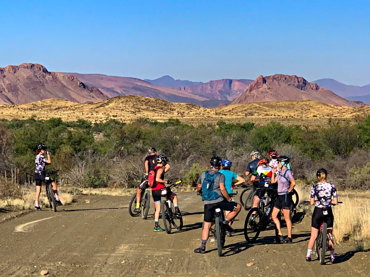 A group of cyclists taking a break on a dirt trail in a desert landscape with mountains in the background.