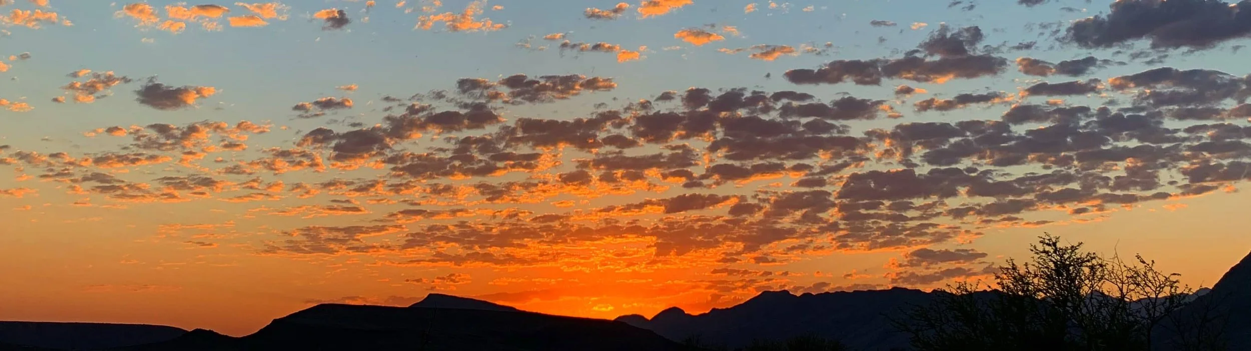 A sunset over mountains with a sky filled with scattered clouds illuminated in orange and yellow hues.
