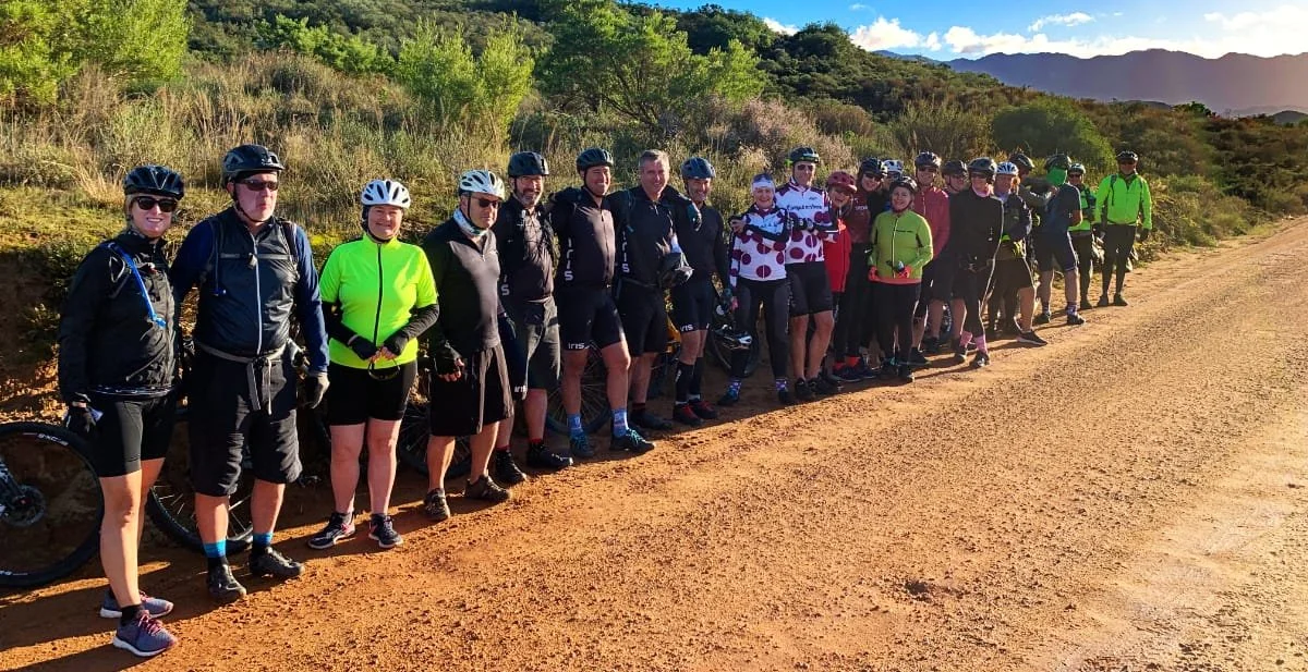 Group of cyclists standing along a dirt road with green trees and hills in the background.