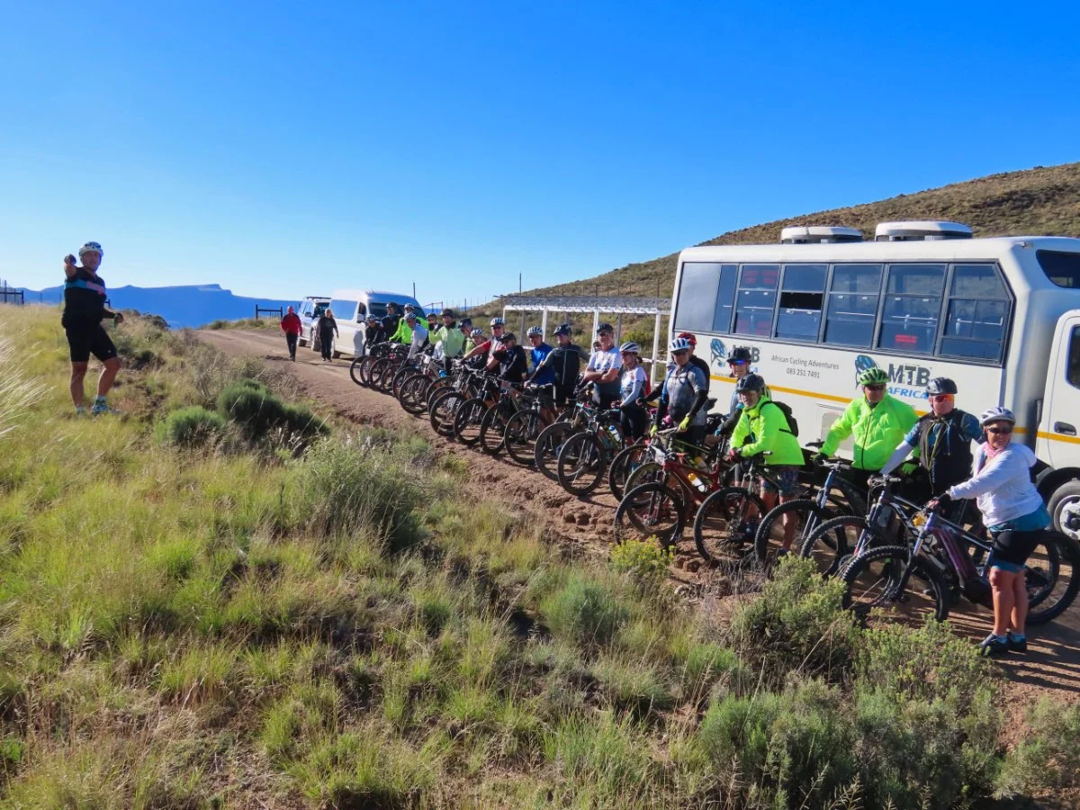 Group of people with bicycles stopped on a dirt trail in a mountainous area, with support vehicles nearby, under a clear blue sky.