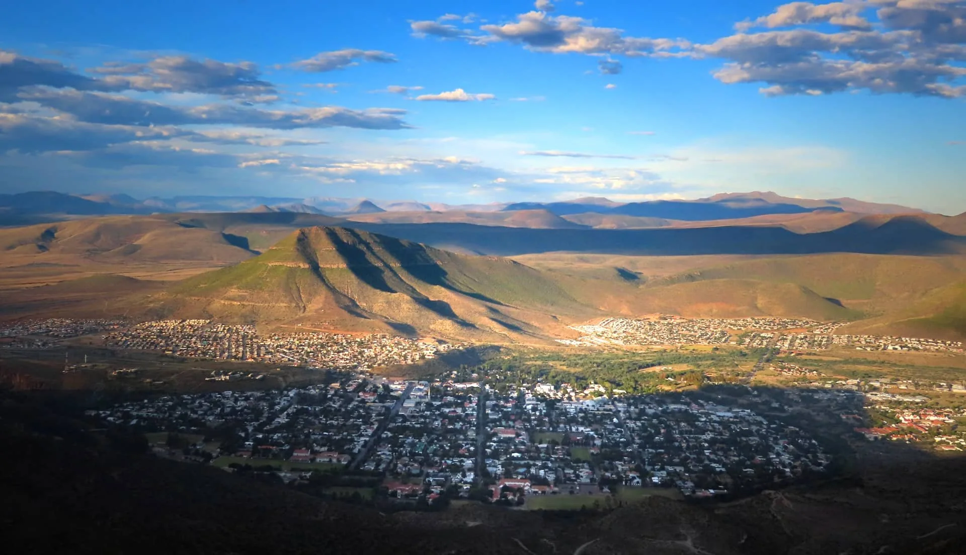 A mountainous landscape with a town at the base, surrounded by rolling hills and a partly cloudy sky.