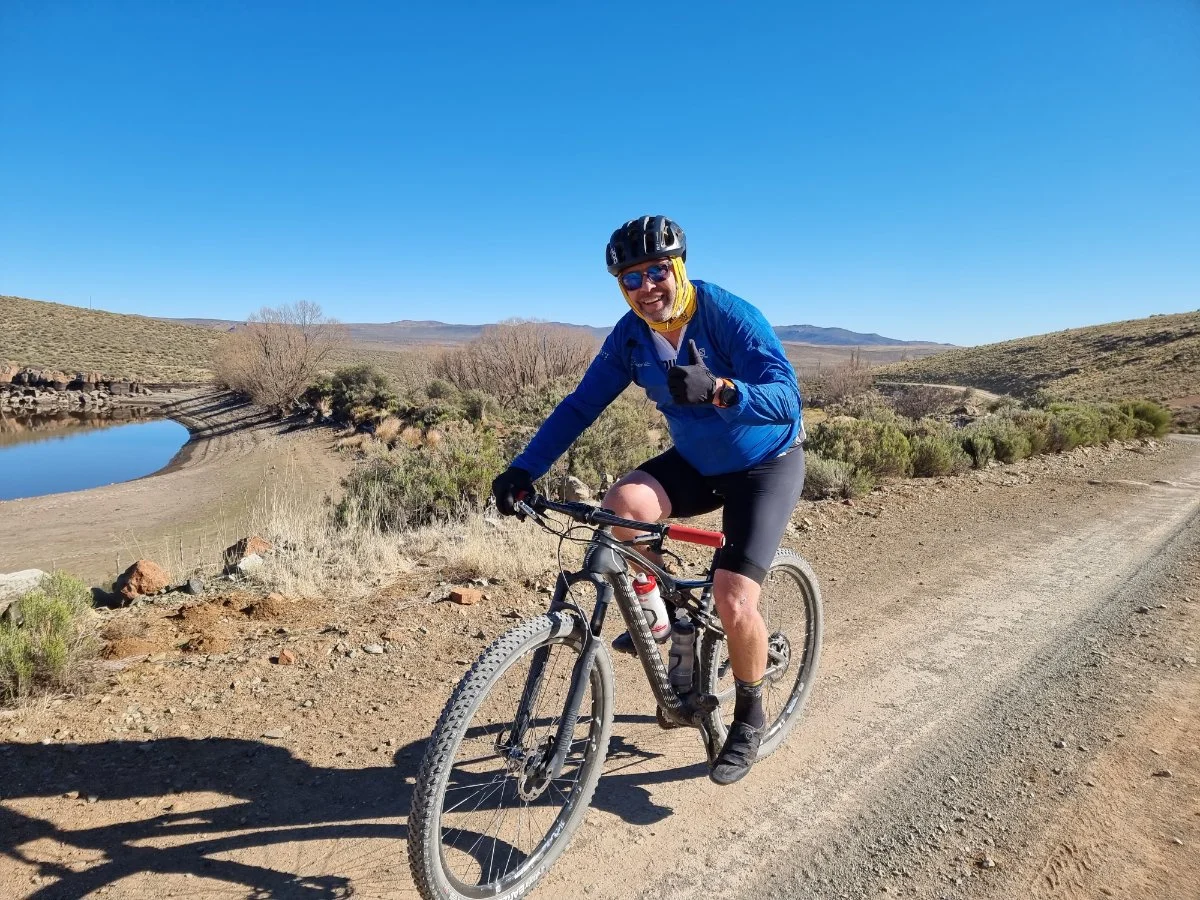 Man in blue jacket and helmet riding a mountain bike on a dirt trail in a desert landscape with sparse shrubs, a small water body, and mountains in the background under a clear blue sky.