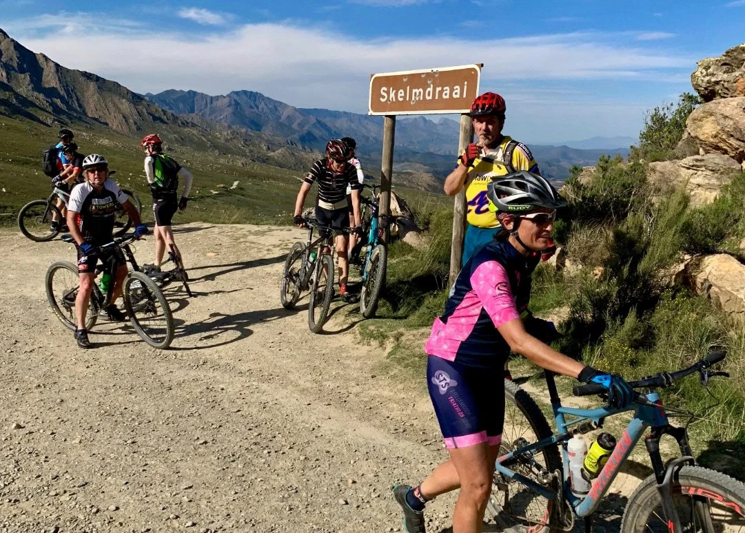Group of mountain bikers resting on a dirt trail in Skelmdraai with mountains and blue sky in the background.