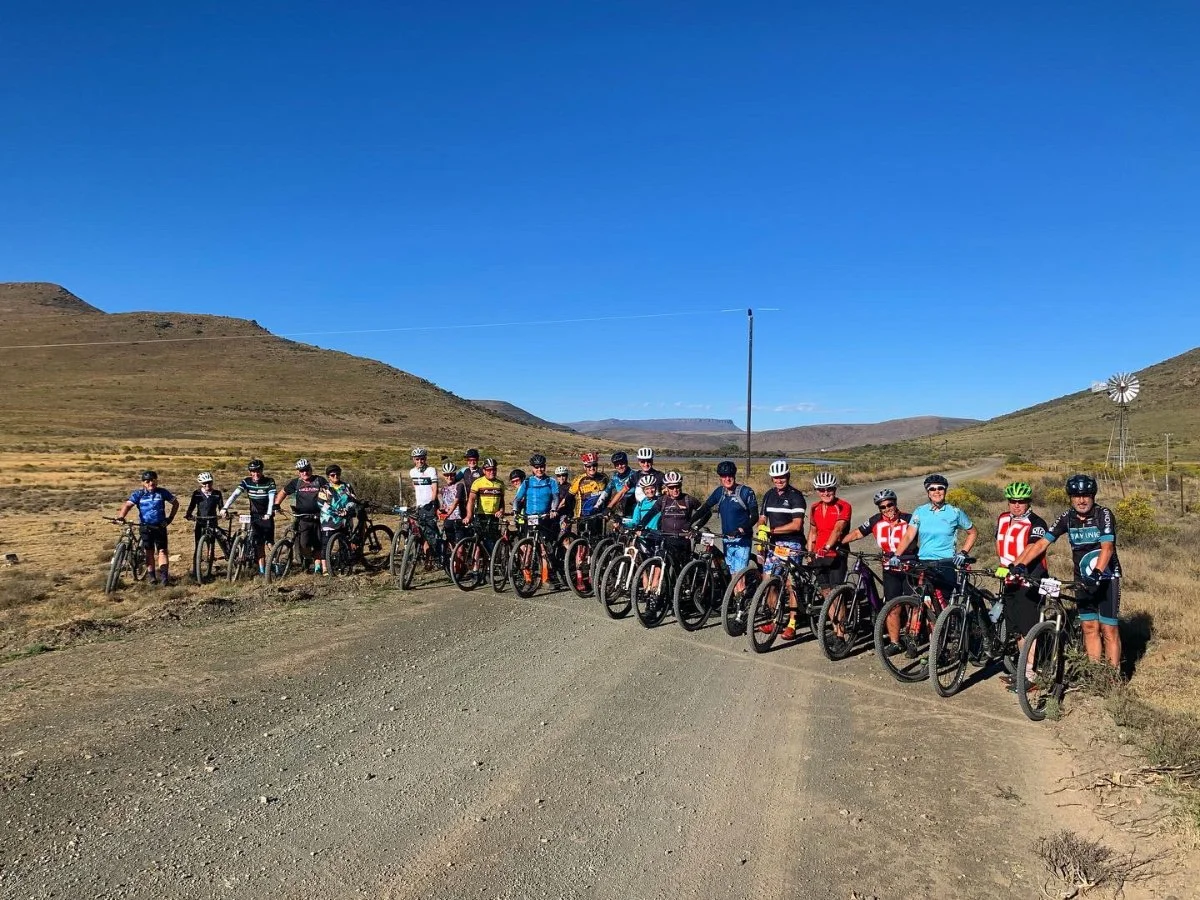 Group of 15 cyclists with helmets and bikes standing on a dirt road in a hilly, arid landscape under a clear blue sky.
