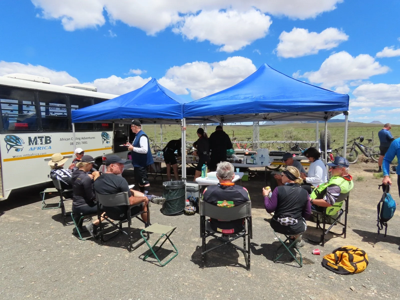 Group of people sitting and standing under blue canopy tents at an outdoor event on a sunny day with blue sky and clouds, bicycles and a bus nearby.