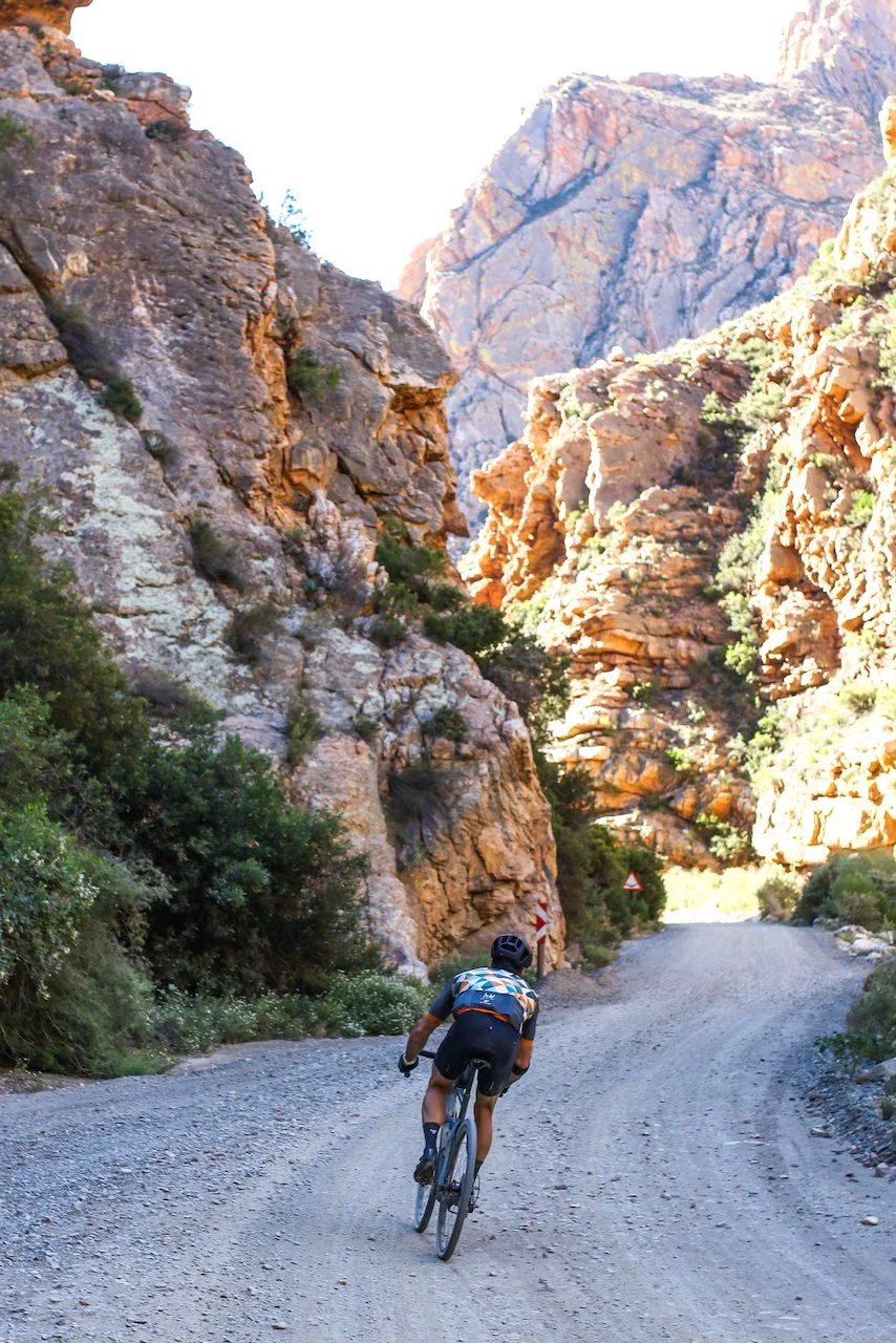 A cyclist riding uphill on a gravel road through a rocky canyon with tall cliffs and sparse vegetation.