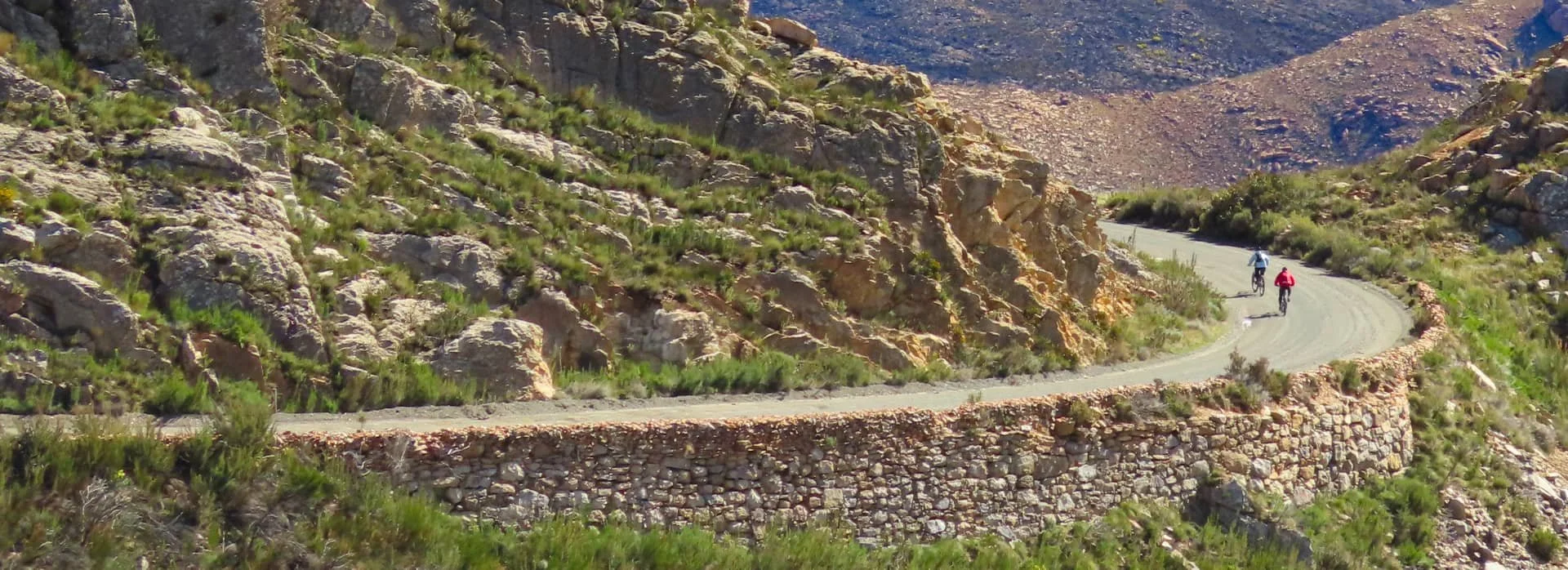 Two cyclists riding bicycles on a winding mountain road with rocky terrain and green shrubs.