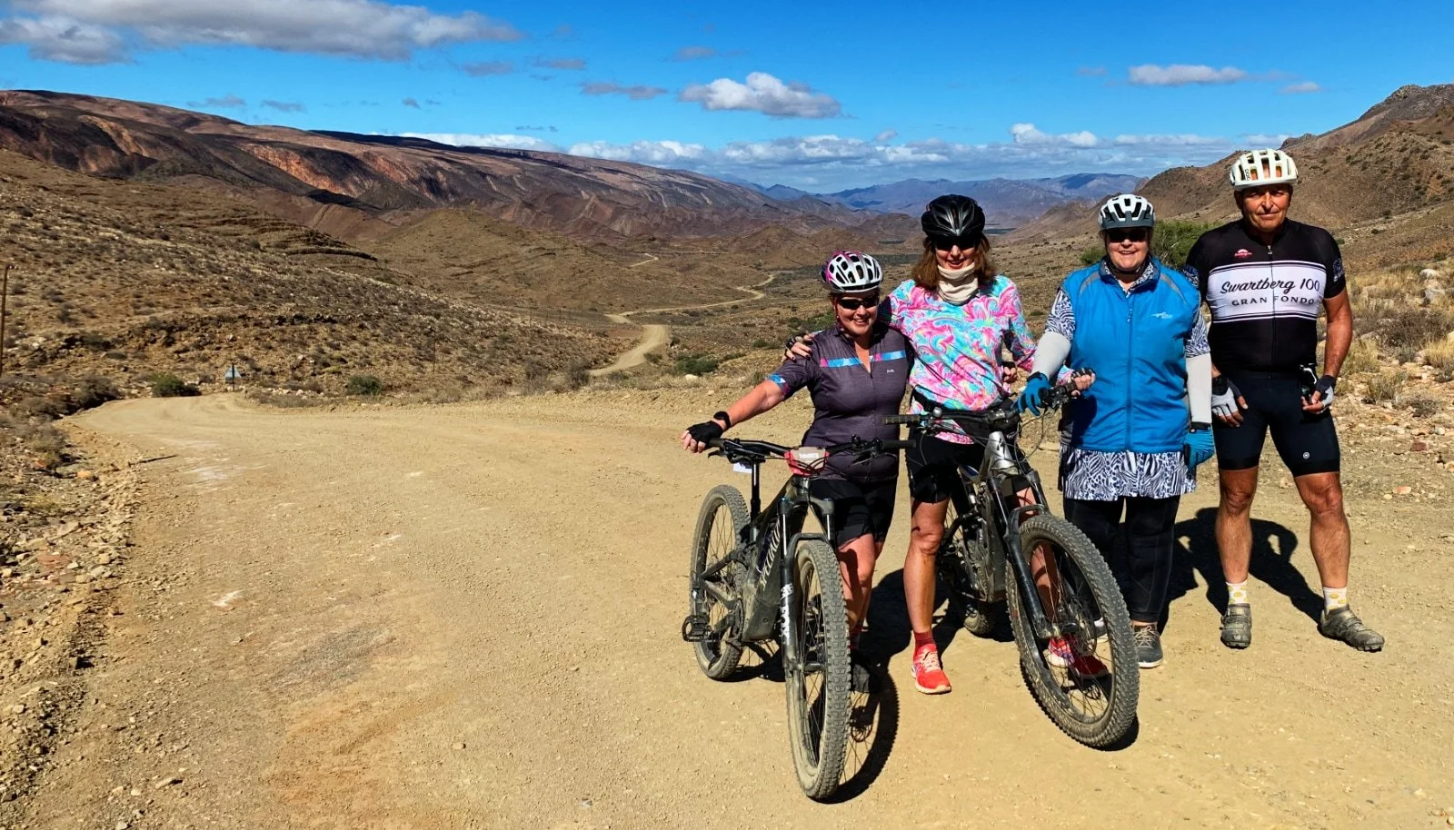 Four people in cycling gear standing on a dirt trail with mountains and blue sky in the background, smiling with two holding bicycles.