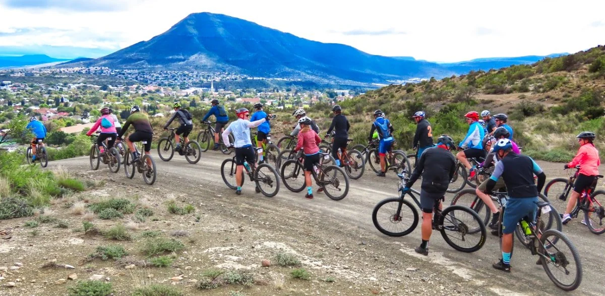 Group of cyclists riding on a dirt trail in a hilly area with a mountain and a town in the background.