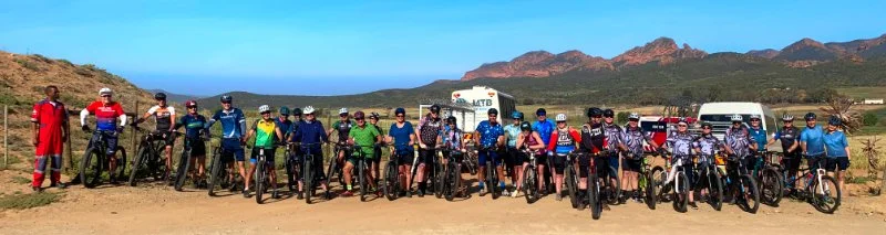 Group of people with mountain bikes standing outdoors on a dirt path, with mountains and blue sky in the background.