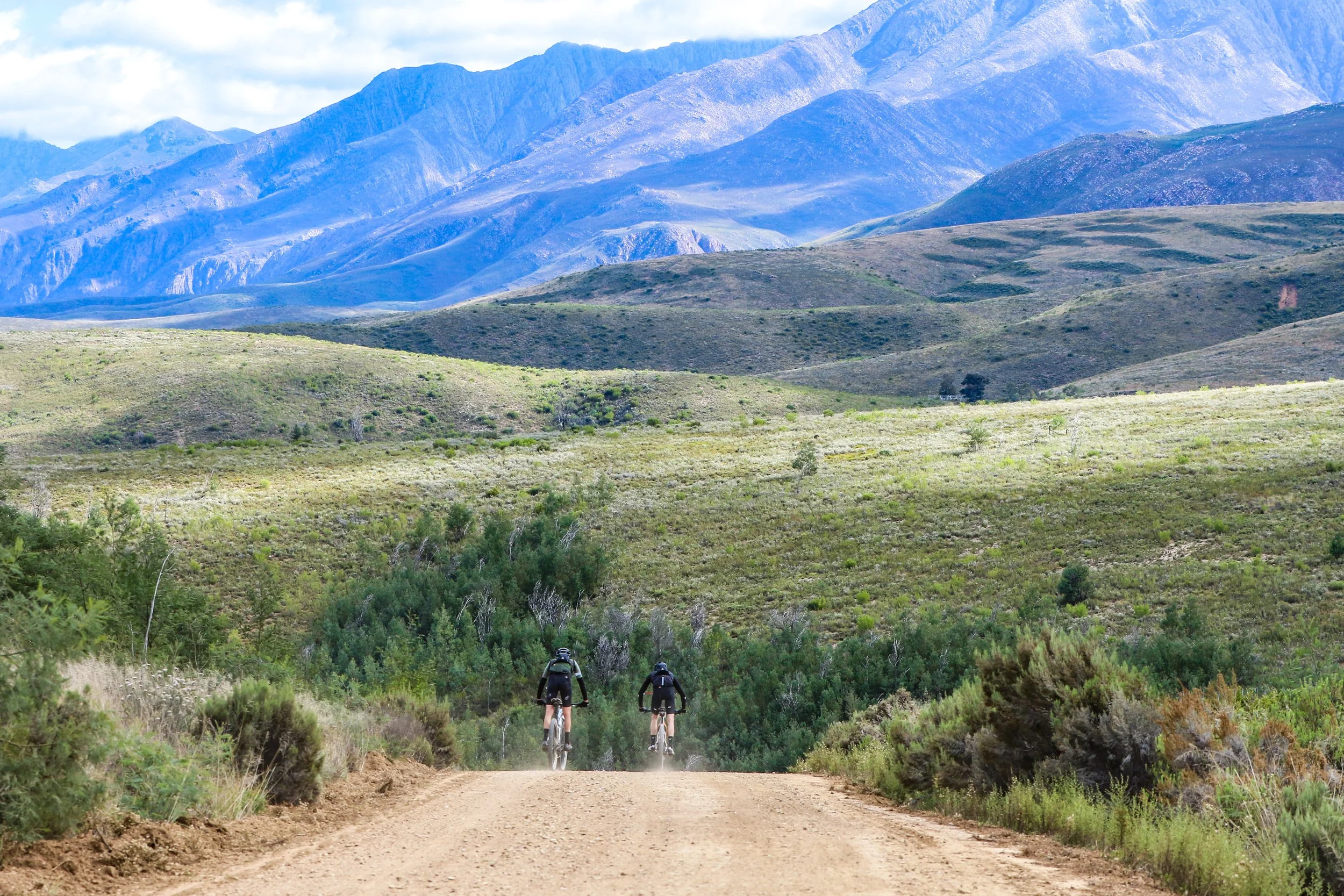 Two cyclists riding a dirt trail in a green valley with hills and mountains in the background.