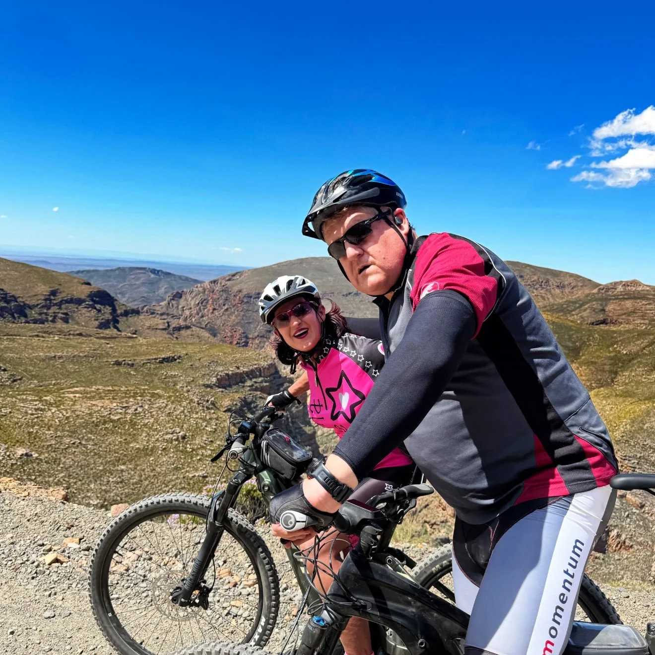 Two people mountain biking on a trail in a mountainous landscape under a clear blue sky