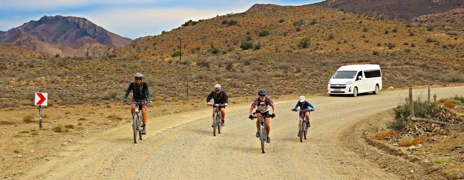 Four people are riding mountain bikes on a dirt road in a desert landscape with mountains in the background. There is a white van parked on the side of the road and a red and white arrow sign indicating a curve.