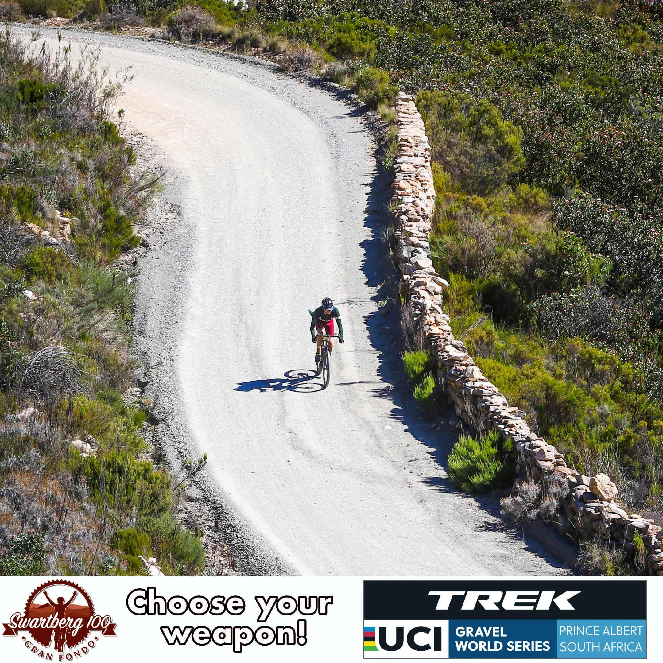 A cyclist riding on a curving gravel road with a stone wall on the right and greenery on both sides.