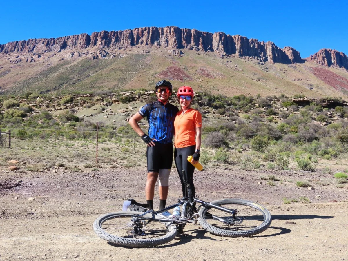 Two cyclists, a man and a woman, stand together on a dirt trail in a desert landscape with mountains in the background. They are wearing helmets, sunglasses, and cycling gear, with a mountain bike lying on the ground in front of them.