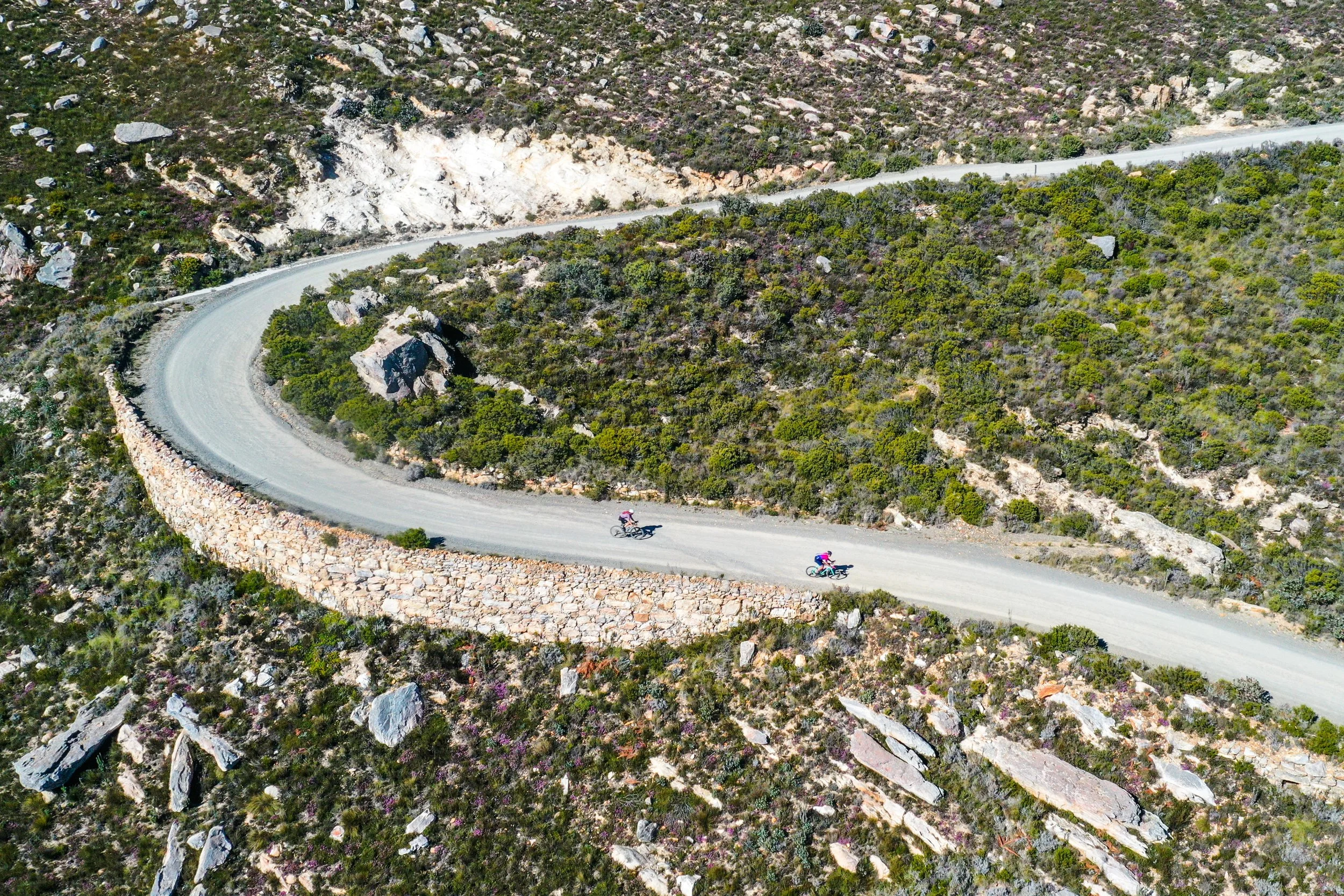 Two cyclists riding on a winding mountain road surrounded by rocky terrain and green shrubbery from an aerial view.