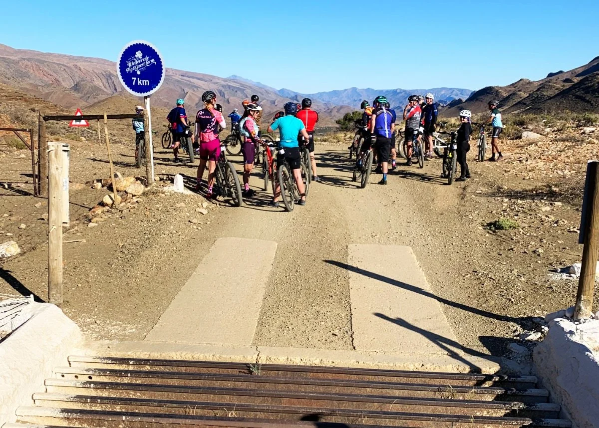 Group of cyclists with helmets resting on a dirt trail in a mountainous desert landscape, near a sign indicating a 7 km distance.
