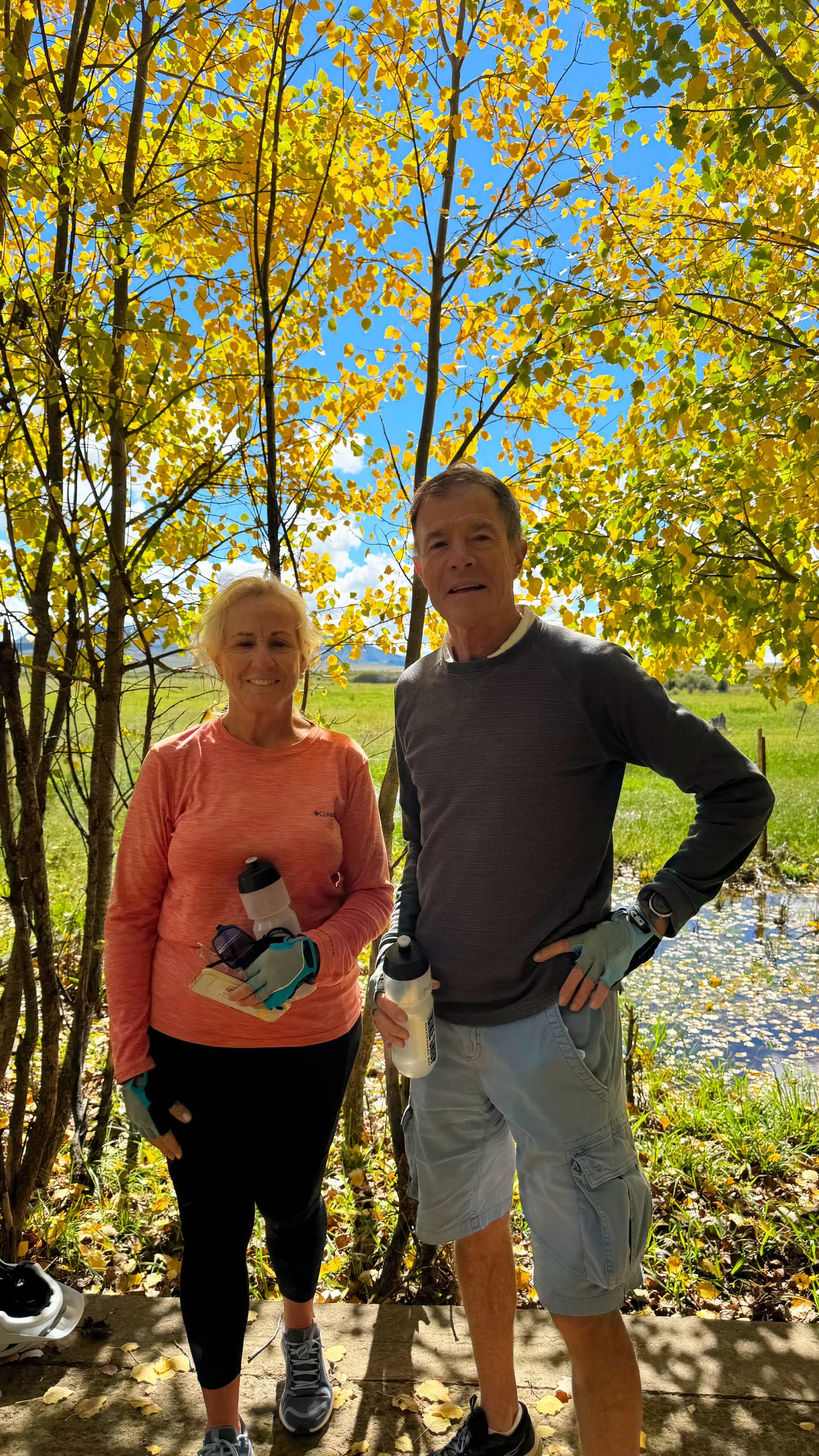 Older man and woman standing outdoors on a trail, holding water bottles, with trees with yellow leaves and a pond in the background.