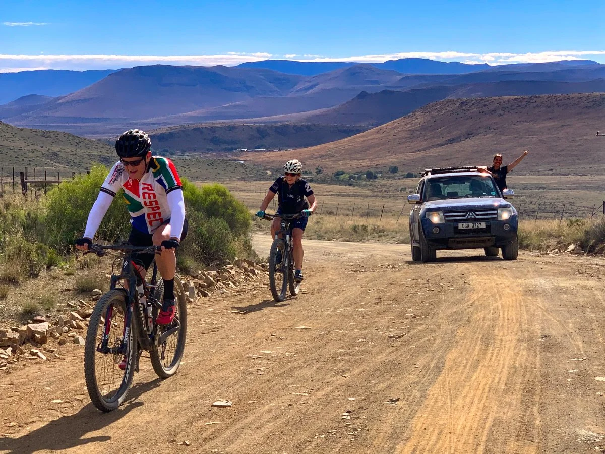 Two people riding mountain bikes on a dirt trail in a desert landscape with mountains in the background, and a person in a vehicle waving.