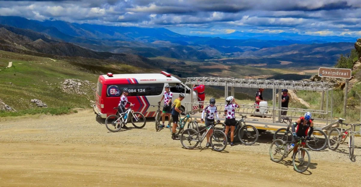 Cyclists and a rescue vehicle with a ladder on a mountain trail with a scenic valley and mountains in the background.