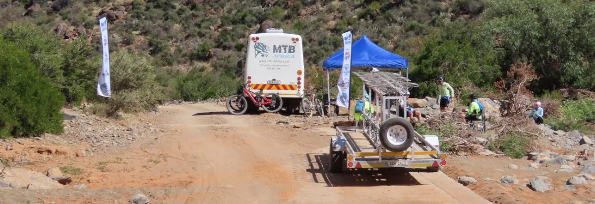 People participating in a mountain biking event on a dirt trail, with a support van, a trailer, and a blue canopy tent nearby in a desert landscape.