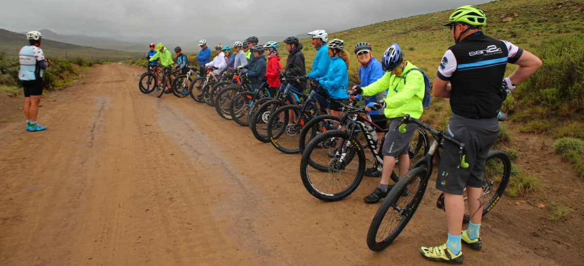 Group of cyclists with helmets and gear lining up on a dirt trail outdoors, with a grassy hillside and cloudy sky in the background.