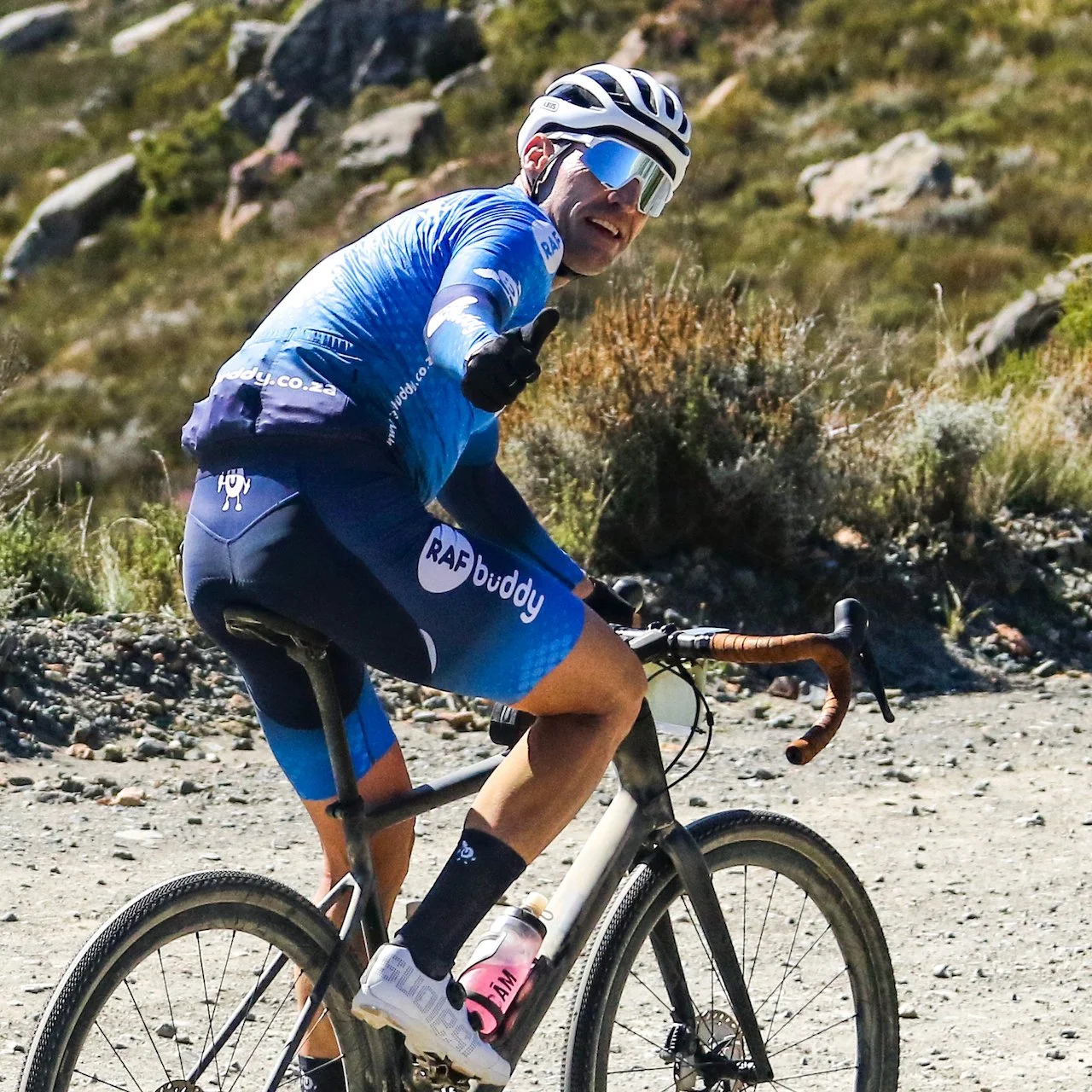 Cyclist wearing sunglasses and a blue jersey riding a mountain bike on a dirt trail in a natural landscape.