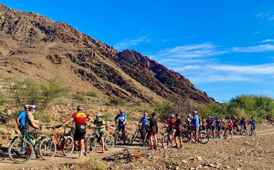 A group of cyclists taking a break on a dirt trail in a mountainous desert landscape under a bright blue sky.
