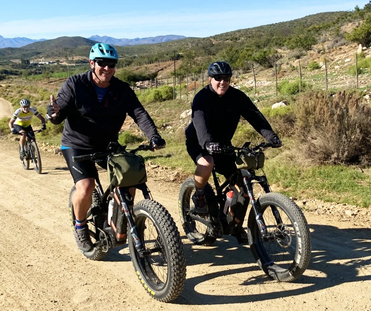 Group of people riding mountain bikes on a dirt trail in a rural area with hills and sparse vegetation under a clear blue sky.