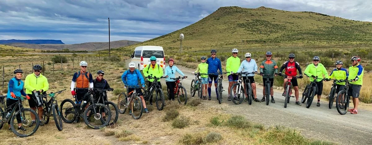 Group of 13 people standing with bicycles outdoors in a desert landscape, with hills in the background and a cloudy sky.