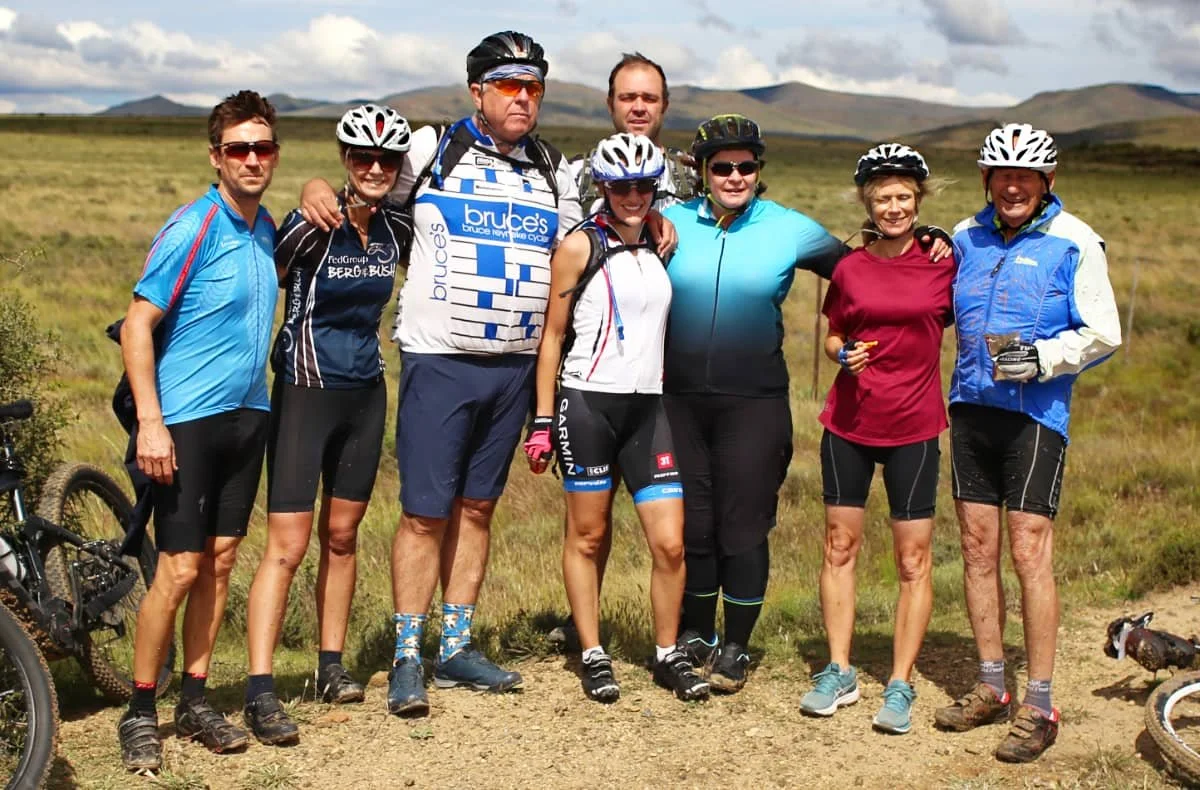 Group of eight people in cycling gear posing outdoors in a grassy landscape with mountains in the background.